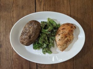 Spicy brown suger baked chicken with a baked potato and roasted broccoli on a white oval plate. Rustic farmhouse table. Cooking chicken from scratch. Homemade dinner. Old fashioned cooking at home. Cozy food.